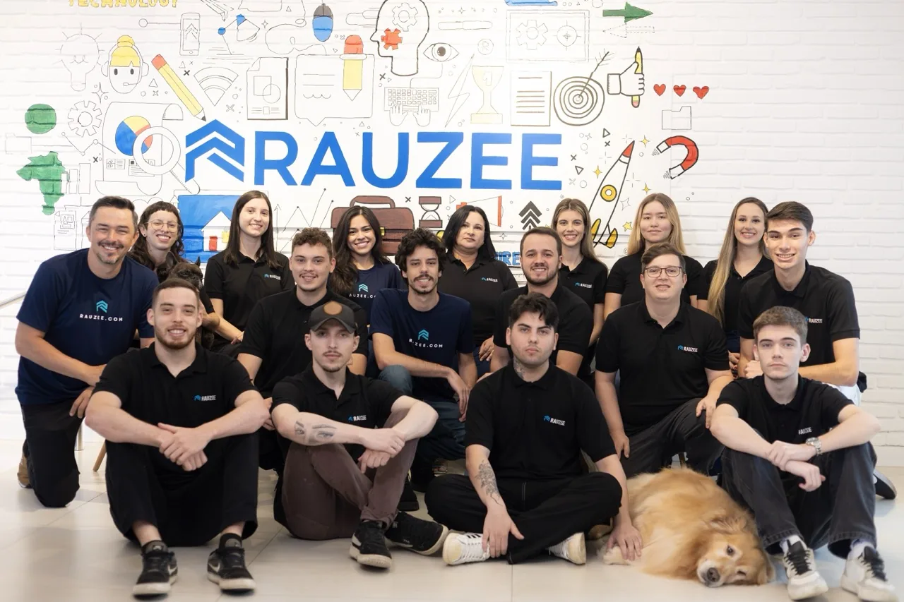 Time da Rauzee reunido no escritório, de camiseta da marca, em frente ao mural com o logo